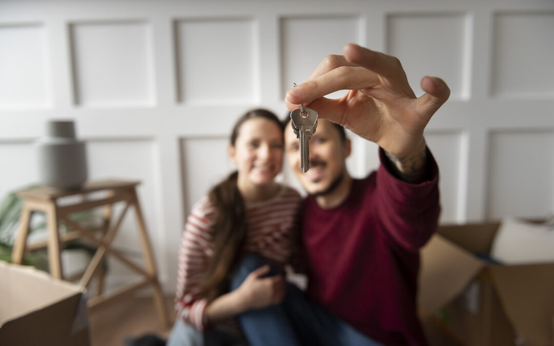 pareja joven feliz mostrando la llave de su nueva casa con cajas de mudanza al fondo