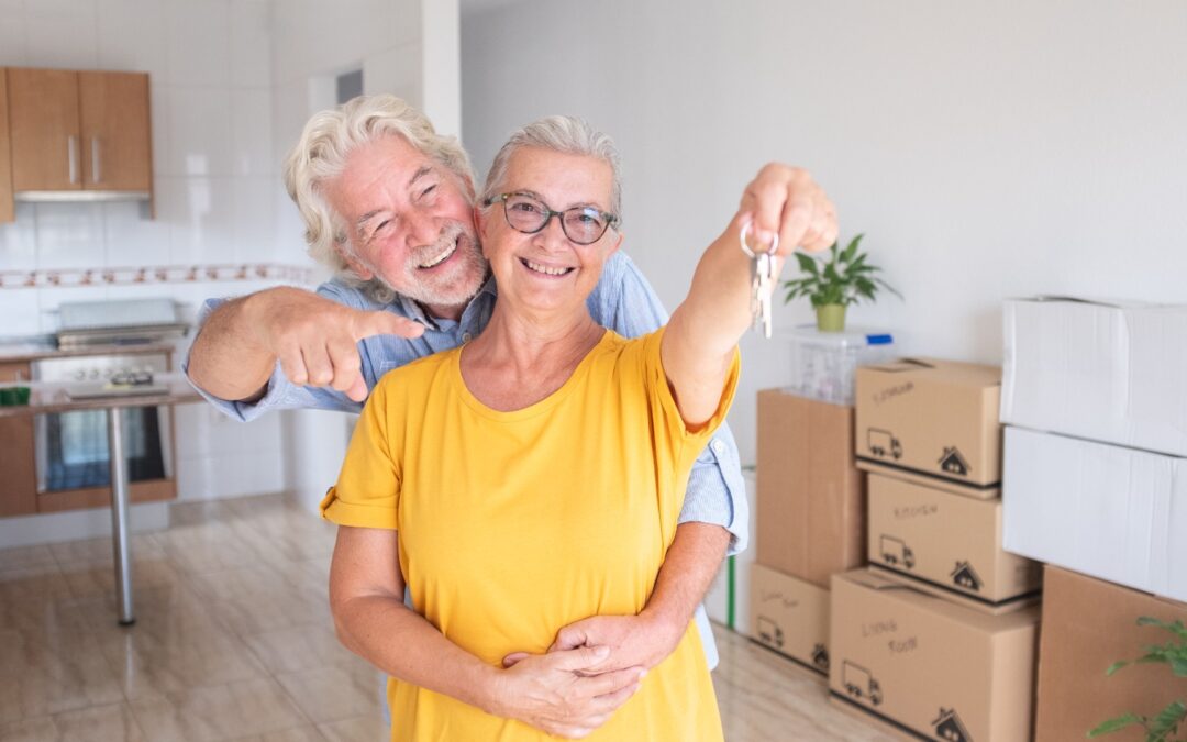 Feliz pareja senior sonriendo con llaves de su nuevo hogar, rodeados de cajas de mudanza en un luminoso salón, ilustrando el exitoso proceso de reubicación para retirados en la Axarquía.